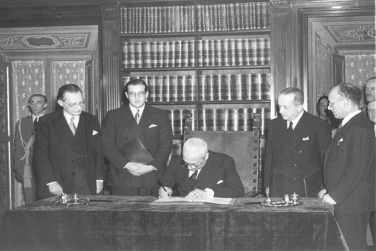 Enrico De Nicola firma la Costituzione italiana nel 1947, seduto a una scrivania circondato da figure istituzionali.