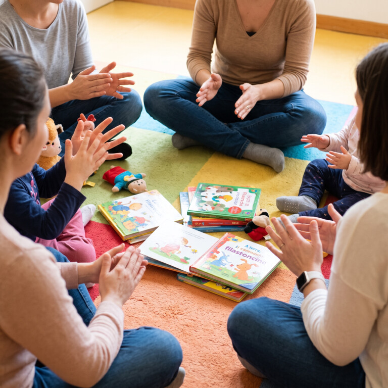 Inquadratura dall'alto e di spalle di mamme e bambini piccoli seduti in cerchio su un tappeto colorato, intenti a battere le mani e a mimare canzoncine insieme, con libri illustrati di filastrocche e piccoli giochi di stoffa sparsi al centro