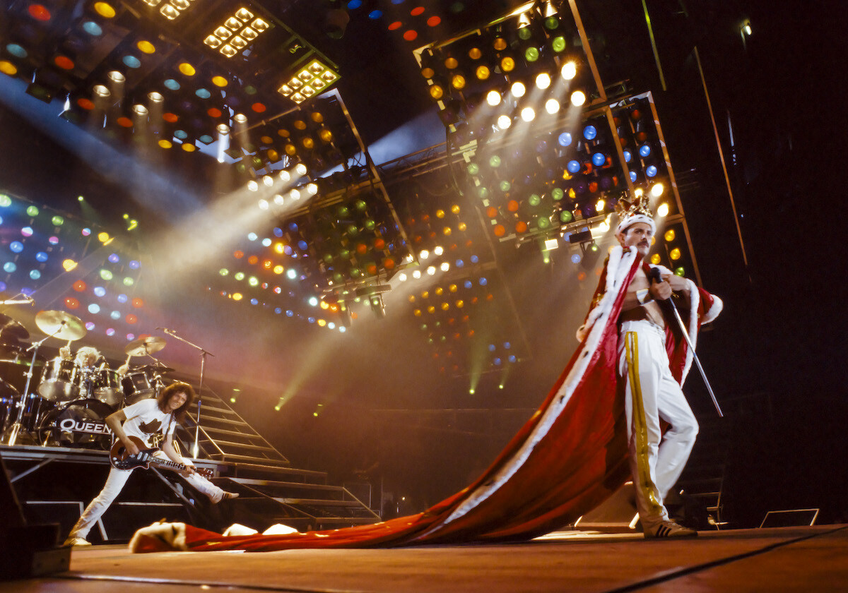 Freddie Mercury con corona reale e lungo mantello rosso sul palco durante un concerto dei Queen nel 1986, con Brian May e luci colorate sullo sfondo.