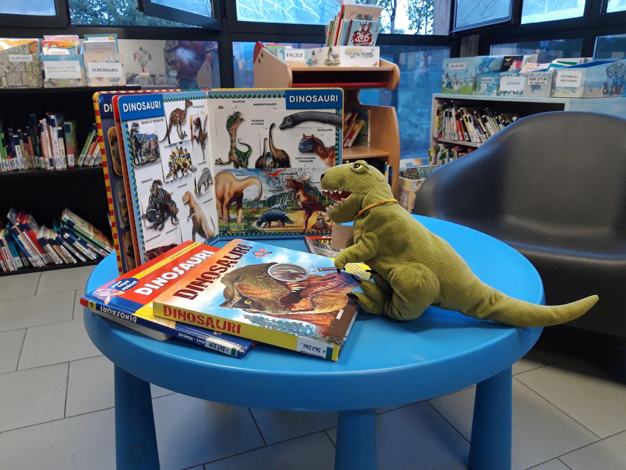 foto di un pupazzo di peluche a forma di dinosauro verde posizionato su un tavolino azzurro per bambini all'interno di una biblioteca; attorno al peluche ci sono diversi libri illustrati sui dinosauri, uno dei quali aperto in verticale, con scaffali ricchi di libri per l'infanzia e una poltrona sullo sfondo