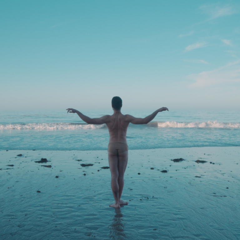 Foto di un danzatore di spalle a torso nudo, con le braccia alzate che disegnano una posa, di fronte al mare