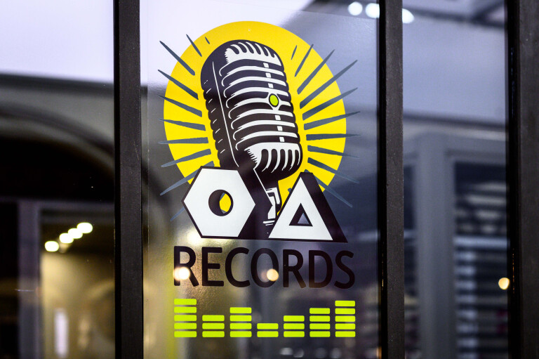foto del logo "OA Records" applicato su una vetrata; l'adesivo mostra un microfono vintage in stile anni '50 racchiuso in un cerchio giallo raggiante, sovrapposto alle lettere geometriche O e A, con la scritta nera "RECORDS" e un grafico di equalizzatore verde acido in basso.