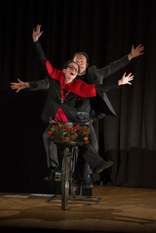 Due attori in costume sorridono e posano con gioia a braccia aperte su una bicicletta in tandem, su un palco con uno sfondo di tende scure.