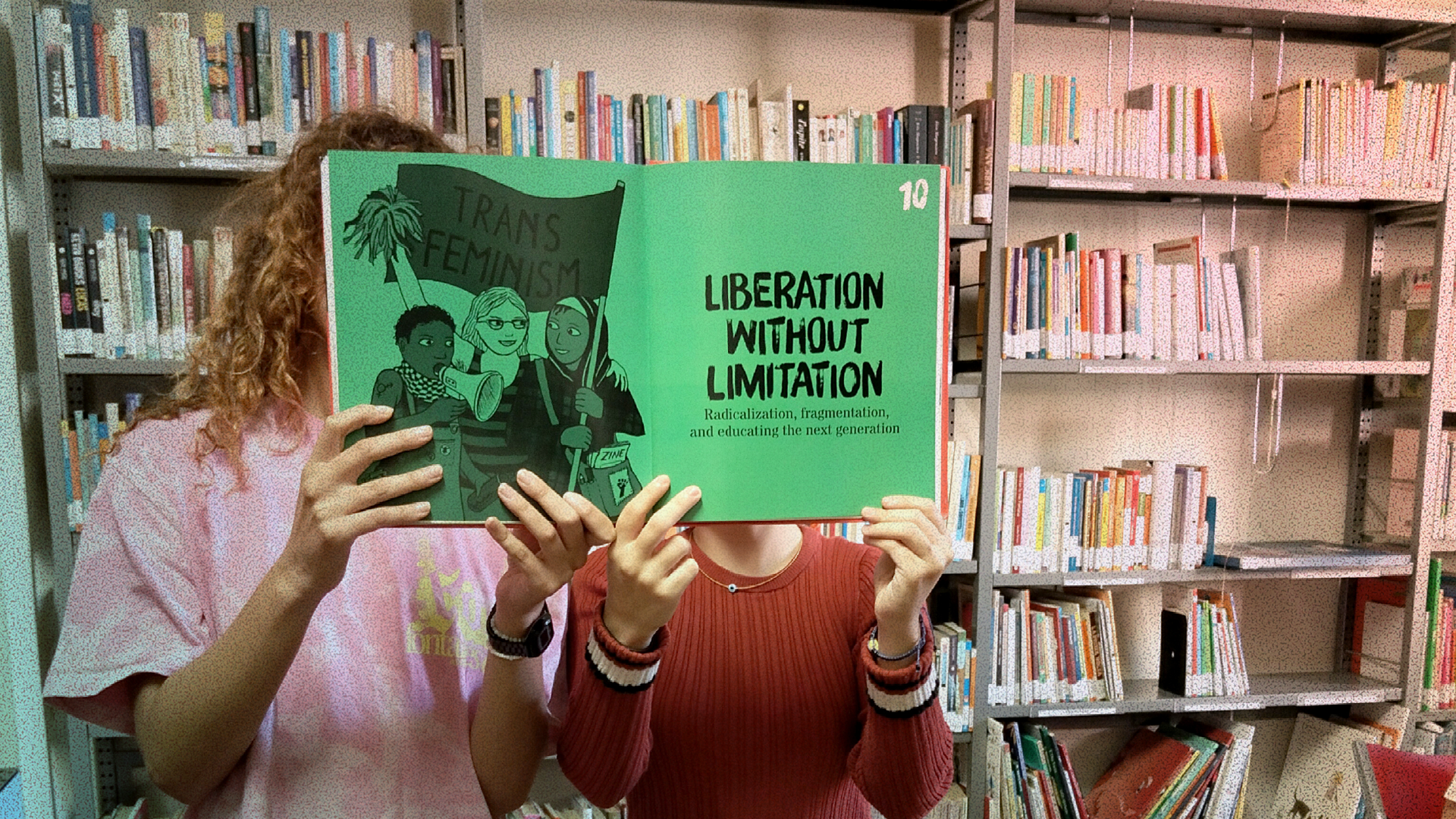 Due persone all'interno di una biblioteca tengono aperto un libro verde con la scritta 'TRANS FEMINISM' e 'LIBERATION WITHOUT LIMITATION' coprendo i propri volti