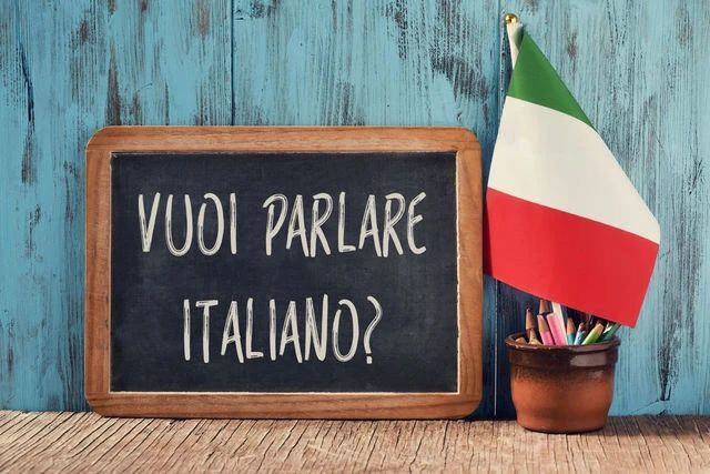 Una lavagnetta in cornice di legno appoggiata su una superficie rustica, con la scritta in gesso bianco "VUOI PARLARE ITALIANO?". A destra della lavagna c'è un piccolo vaso in ceramica con matite e una bandierina tricolore italiana. Lo sfondo è composto da assi di legno verniciate di azzurro.