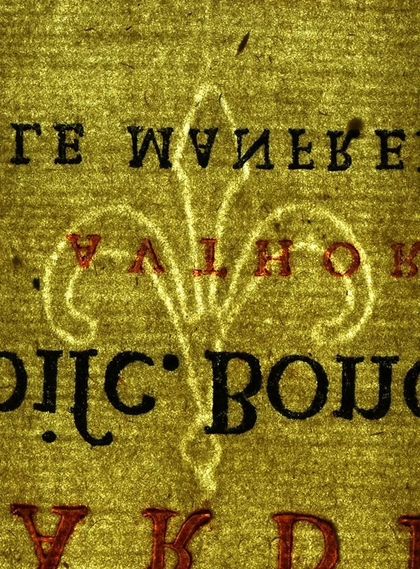 foto di una pagina di carta antica in filigrana illuminata da dietro, di colore giallo dorato, con impresso al centro un giglio stilizzato in chiaro; sopra e sotto la filigrana sono visibili lettere e parole stampate a specchio sia in nero che in rosso.