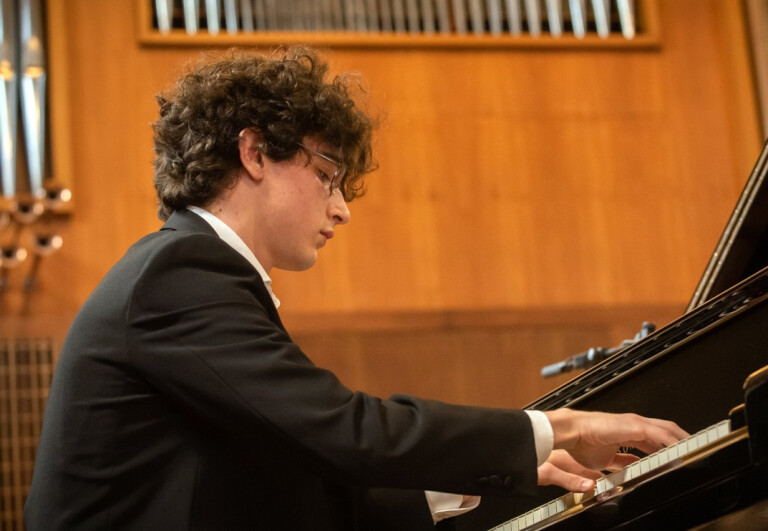 Il giovane musicista Tommaso Franchini, giacca nera e camicia bianca, è seduto al pianoforte e sta suonando durante un'esibizione. 
