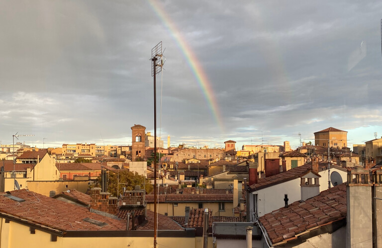 panorama della città di Bologna al tramonto, con distese di tetti in terracotta e camini, sotto un cielo nuvoloso drammatico e illuminato, da cui spunta un vivido arcobaleno diagonale