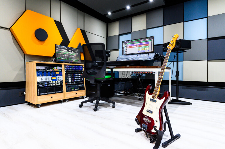 foto all'interno di una moderna sala di registrazione e missaggio; in primo piano a destra un basso elettrico su supporto da terra, al centro una scrivania con sedia ergonomica, console di missaggio e monitor con software audio aperto, affiancata a sinistra da moduli rack con processori professionali su pareti insonorizzate a pannelli modulari blu, grigi e arancioni