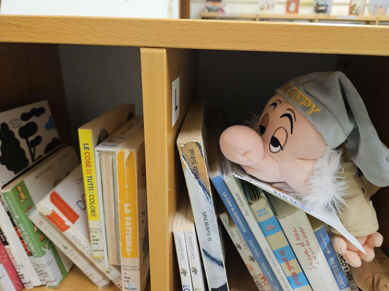 foto ravvicinata di due scomparti di uno scaffale di legno pieno di libri per bambini; a destra, un pupazzo del nano Pisolo della Disney sbuca tra i libri con lo sguardo rivolto in avanti; si distinguono chiaramente i dorsi di diversi libri con titoli leggibili 