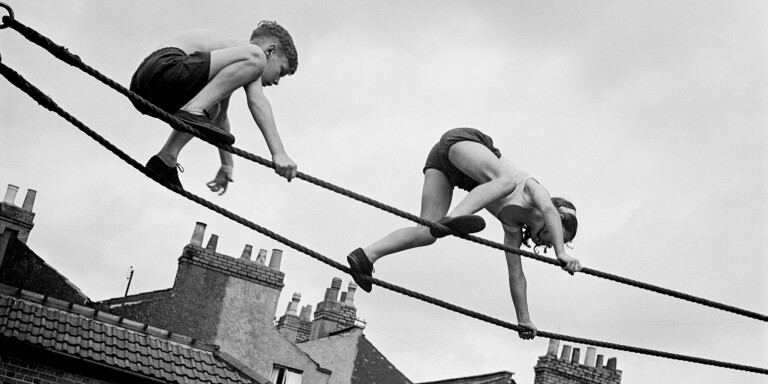 Foto in bianco e nero di Edith Tudor-Hart: due bambini a quattro zampe percorrono in equilibrio due corde tese sopra i tetti di una città.