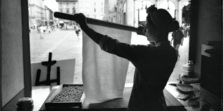 Sfoglina solleva una sottile sfoglia di pasta fresca con il matterello davanti alla vetrina del laboratorio