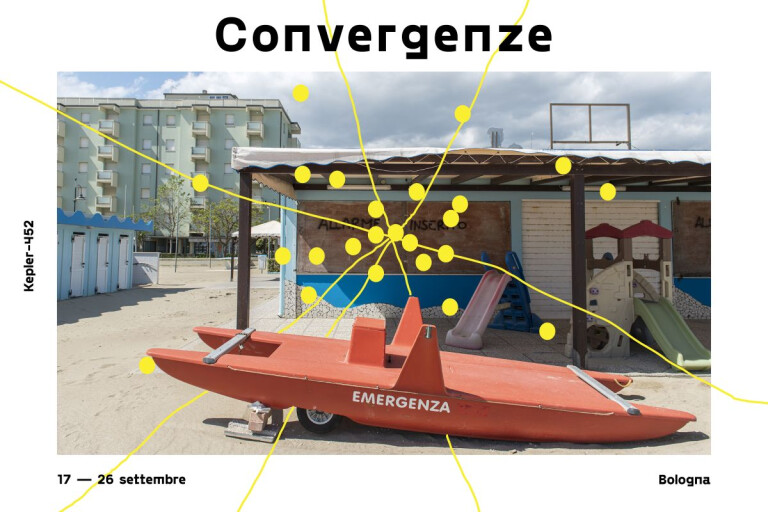 Immagine tratta dalla locandina promozionale dell'evento "Convergenze": la fotografia mostra uno scorcio di una spiaggia italiana fuori stagione o deserta. In primo piano sulla sabbia c'è un pattino di salvataggio (moscone) di colore rosso con la scritta bianca "EMERGENZA" sulla fiancata. Sullo sfondo si notano le strutture di uno stabilimento balneare chiuso, con serrande abbassate, piccoli scivoli di plastica per bambini e cabine azzurre sulla sinistra. Più lontano, dietro lo stabilimento, svetta un condominio o hotel di colore verde chiaro sotto un cielo parzialmente nuvoloso. Una rete di linee e cerchi gialli si sovrappone all'immagine, convergendo in un punto centrale denso di nodi posizionato sopra la struttura dello stabilimento balneare.