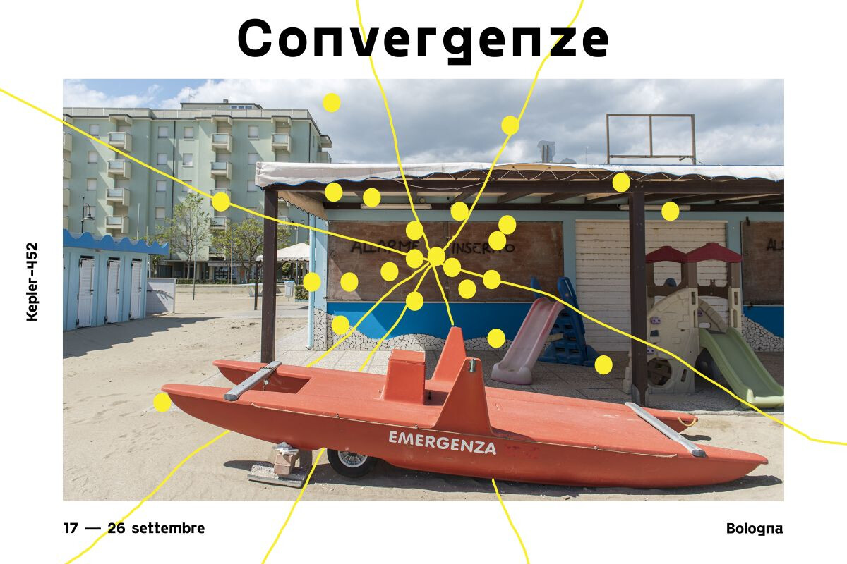 Immagine tratta dalla locandina promozionale dell'evento "Convergenze": la fotografia mostra uno scorcio di una spiaggia italiana fuori stagione o deserta. In primo piano sulla sabbia c'è un pattino di salvataggio (moscone) di colore rosso con la scritta bianca "EMERGENZA" sulla fiancata. Sullo sfondo si notano le strutture di uno stabilimento balneare chiuso, con serrande abbassate, piccoli scivoli di plastica per bambini e cabine azzurre sulla sinistra. Più lontano, dietro lo stabilimento, svetta un condominio o hotel di colore verde chiaro sotto un cielo parzialmente nuvoloso. Una rete di linee e cerchi gialli si sovrappone all'immagine, convergendo in un punto centrale denso di nodi posizionato sopra la struttura dello stabilimento balneare.
